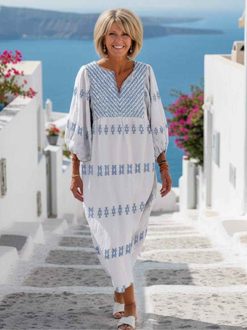 Woman wearing a white embroidered v-neck maxi dress with intricate blue stitching and long sleeves, flowing full-length silhouette