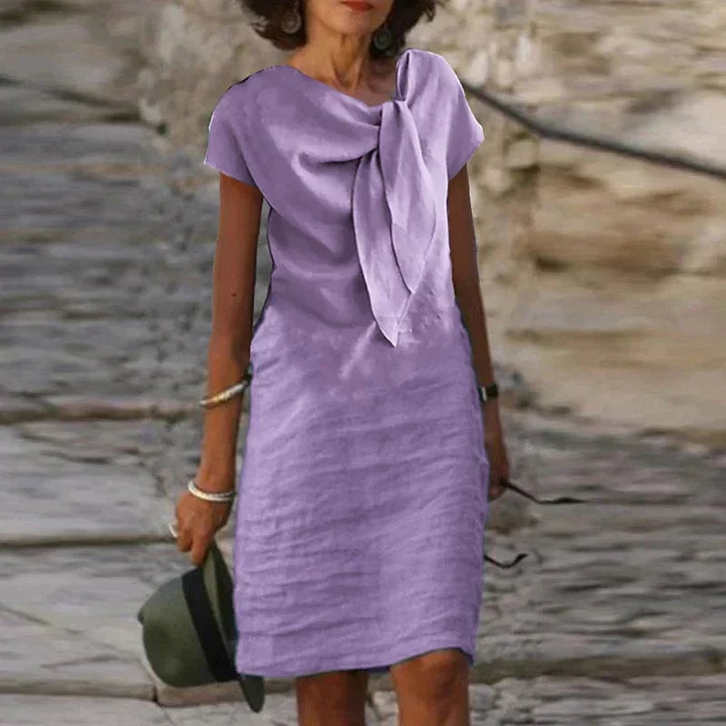 Woman wearing the Celeste bow neck short sleeve midi dress in purple, featuring a draped bow neckline and knee-length hem