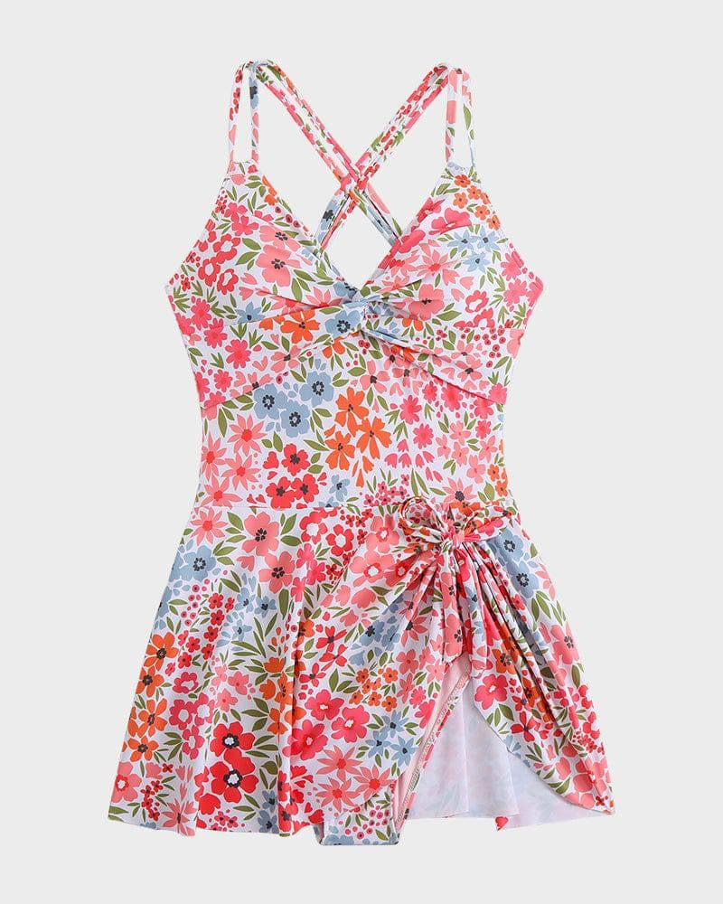 Model wearing orange floral swim dress with skirted overlay, standing near the water